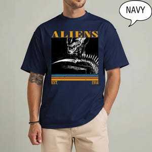 May include: Navy blue t-shirt featuring a graphic of an alien creature with the word "ALIENS" in gold lettering. Below the image are the words "EST. 1986" in a retro style. The shirt is a classic crew neck.