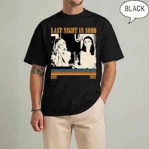 May include: Black t-shirt featuring a graphic from the movie "Last Night in Soho." The design includes two figures, the text "LAST NIGHT IN SOHO," and the year "2021." The shirt has a retro style with horizontal stripes in blue, orange, and yellow.