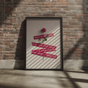 Könnte beinhalten: Ein gerahmter Kunstdruck mit einer einzelnen roten Rose, die auf einem hellen Hintergrund zentriert ist. Drei rote Klebestreifen sind horizontal über die Rose gelegt, jeweils bedruckt mit dem weißen Text: WHAT IS YOUR LOVE SONG?