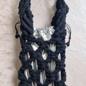 May include: A small bag featuring a metallic silver inner pouch with a drawstring closure, encased in a black macrame net overlay. The bag includes a thick, braided black cord handle that extends from the top of the knotted net structure.