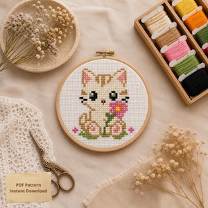 Pode incluir: Um bastidor de bordado em ponto cruz com um gato fofo segurando uma flor rosa. O gato é bege com listras marrons e olhos verdes. O texto "PDF Pattern Instant Download" está no canto inferior esquerdo. Uma caixa de madeira com carretéis de linha coloridos está ao fundo.