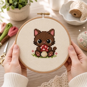 Pode incluir: Um bastidor de bordado em ponto cruz com um gato castanho segurando um cogumelo vermelho e branco. O gato está sentado na grama verde com pequenas flores cor-de-rosa. O bordado está num bastidor de madeira.