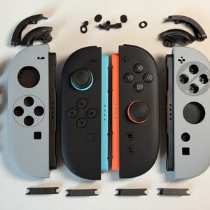 May include: Disassembled grey and black game controllers with various coloured buttons and black components. The controllers are separated, showing internal parts and buttons. The controllers have blue and orange accents.
