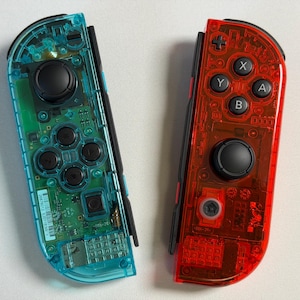 May include: Two transparent game controllers, one teal and one red, are shown. The teal controller has black buttons and a black joystick. The red controller has black buttons and a black joystick. Internal components are visible on both controllers.