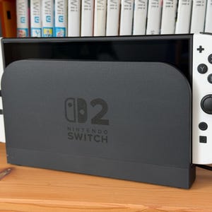 May include: A black Nintendo Switch dock with the Nintendo Switch logo and the number 2 printed on the front. The console is docked with white Joy-Con controllers attached to the sides, featuring black buttons and joysticks on a white frame.