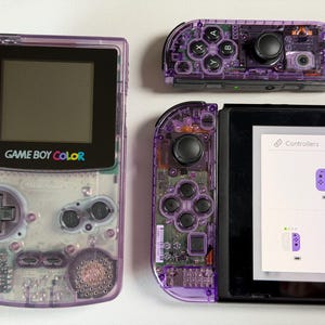 May include: A transparent purple Game Boy Color console and two Nintendo Switch controllers. The Game Boy Color has a black screen and the words "GAME BOY COLOR" in white and colour text. The Switch controllers are purple and show internal components.