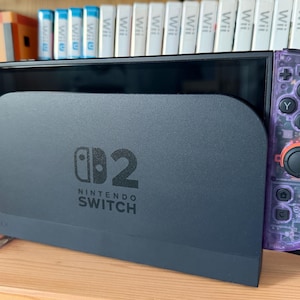 May include: A black Nintendo Switch console docked in a black charging station with the Nintendo Switch logo printed on the front. The console features custom translucent purple Joy-Con controllers with black buttons and a blue joystick on the left.