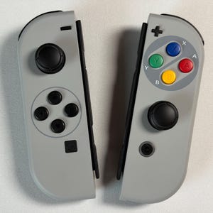 May include: Two grey game controllers, each with a black joystick and buttons. One controller has a directional pad and four black buttons. The other has four coloured buttons: blue, green, red, and yellow, along with a plus-shaped directional button.