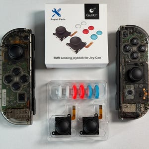 May include: Two transparent black game controllers with visible internal circuitry, a white box labelled Repair Parts GuliKit TMR sensing joystick for Joy-Con, and a plastic tray containing two black joystick modules and six coloured caps.