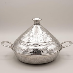 Può includere: Una pentola tagine in metallo argentato martellato con coperchio conico e due manici ad anello. La pentola ha una superficie strutturata e una fascia decorativa attorno alla base del coperchio. Il coperchio è sormontato da un piccolo pomello rotondo.