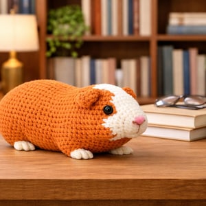 Könnte beinhalten: Ein gehäkeltes Meerschweinchen in Orange- und Weißtönen sitzt auf einer Holzoberfläche. Das handgefertigte Tier hat schwarze Augen und eine rosa Nase. Der Hintergrund umfasst ein Bücherregal, Bücher und eine Lampe.