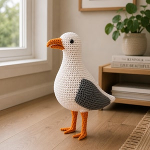 May include: A handcrafted crochet seagull plush toy featuring a white head and body, dark gray wings, and a bright orange beak and feet. The bird has small black bead eyes and is constructed with a visible yarn stitch pattern throughout its body.