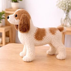 May include: A handcrafted crochet dog figurine in cream and brown. The dog has brown ears and spots, with black eyes and a small tail. The dog is standing on a wooden surface.