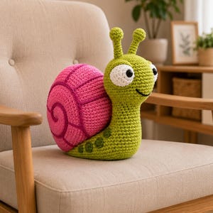 Puede incluir: Un juguete de caracol tejido a ganchillo con un cuerpo verde brillante, manchas verde oscuro y dos antenas. El caracol tiene una concha rosa grande, redonda y texturizada con detalles en espiral rosa oscuro y grandes ojos blancos con pupilas negras.