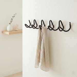 May include: A black metal wall-mounted coat rack with six curved hooks. A beige scarf hangs from one of the hooks. A small white vase with greenery and a spool of twine sit on a wooden shelf to the left.