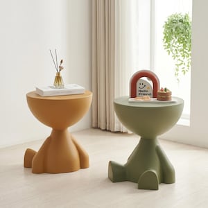 Puede incluir: Dos mesas auxiliares modernas, una amarilla mostaza y otra verde oliva. Cada una tiene una parte superior redondeada y una base con forma de persona sentada con las piernas extendidas. La mesa amarilla tiene un libro y un jarrón. La mesa verde tiene un arco decorativo y un pastel pequeño.