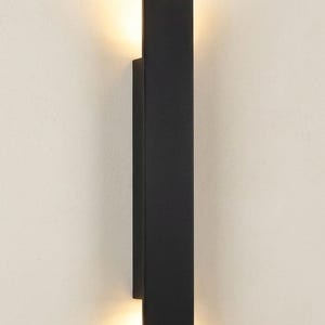 May include: A vertical rectangular wall sconce with a matte black finish. The fixture features an elongated, flat front panel that emits a warm, soft glow from both the top and bottom edges, creating a focused light effect on the wall surface.
