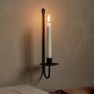 Puede incluir: Un aplique de pared de metal negro con una vela blanca encendida. El aplique tiene una base circular y un brazo largo y delgado que se fija a la pared. La vela es alta y delgada, con una llama parpadeante. La pared es de color neutro.