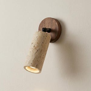 Peut inclure: Un spot mural composé d'une tête cylindrique en pierre travertin beige poreuse, fixée sur une base circulaire en bois brun foncé par une rotule en métal noir, diffusant une lumière chaude depuis l'extrémité ouverte du cylindre en pierre.