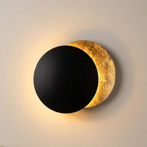 Peut inclure: Une applique murale circulaire composée d'un disque avant noir mat qui chevauche partiellement une plaque arrière circulaire plus grande à la texture dorée. Le luminaire émet une lueur chaude derrière le disque noir, illuminant la surface dorée.