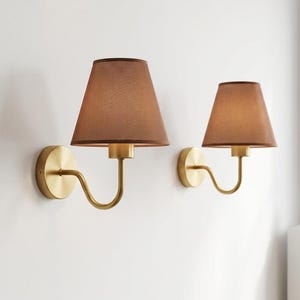 May include: Two wall sconces featuring brown fabric shades and brass-toned metal fixtures. The shades are conical, and the fixtures have a curved arm design. The sconces are mounted on a white wall.