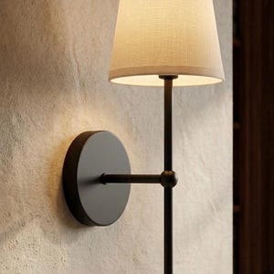 Op de afbeelding: Moderne wandlamp met een zwart metalen frame en een beige stoffen lampenkap. De lamp heeft een ronde basis en een rechte arm. De conische lampenkap geeft een warm licht. Gemonteerd op een getextureerde muur.