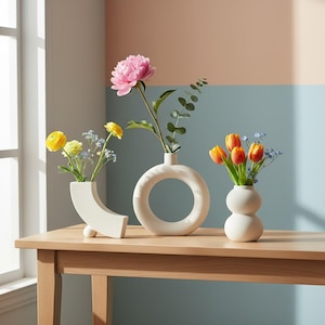 Puede incluir: Tres jarrones de cerámica color crema de varias formas contienen flores coloridas. Una peonía rosa, tulipanes naranjas y flores amarillas se exhiben sobre una mesa de madera clara contra una pared de dos tonos.