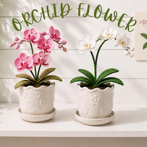 Könnte beinhalten: Zwei gehäkelte Orchideen in weißen Keramiktöpfen. Eine Pflanze hat rosa Blüten, die andere weiße Blüten. Beide haben grüne Blätter und stehen in strukturierten Töpfen mit Herz- und Blumenmustern. Der Text ORCHID FLOWER ist zu sehen.