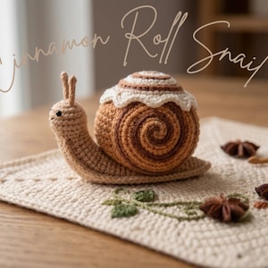 Könnte beinhalten: Gehäkelte Schnecke in Form einer Zimtschnecke, mit braun-cremefarbenem Gehäuse und Körper. Die Schnecke sitzt auf einem cremefarbenen gehäkelten Quadrat. Der Text "Cinnamon Roll Snail" steht darüber.
