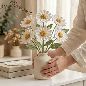 Könnte beinhalten: Ein gehäkelter Gänseblümchenstrauß mit sechs weißen Blüten mit gelben Zentren und grünen Blättern, arrangiert in einer matten beigen Keramikvase. Der Text Daisy Bouquet steht in Schreibschrift in der oberen linken Ecke des Bildes.