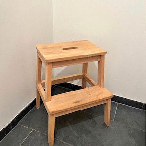 May include: A two-step wooden step stool. The stool is made of light-coloured wood and has a rectangular top with a handhold. The lower step is also rectangular. The stool is designed for reaching higher places.