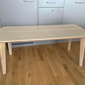 May include: A light-colored wooden coffee table with a rectangular top and four tapered legs. The table is made of light-colored wood and has a simple, modern design. The table is approximately 48 inches long and 24 inches wide.