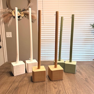 May include: A collection of wooden display stands in various colors. The stands feature a square base and a vertical pole. Colors include white, natural wood, and green. The stands are arranged on a wooden surface.