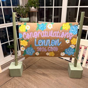 May include: A handmade graduation banner with the words "Congratulations Lennon 2026 Grad" in purple and blue lettering. The banner is decorated with yellow, blue, and white floral accents. The banner is supported by two green wooden posts.