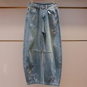 Könnte beinhalten: Hellblaue Jeans aus Denim mit hoher Taille, Knopf- und Reißverschluss sowie weit geschnittenen Beinen. Der Stoff ist mit verschiedenen kleinen, bunten gestickten Aufnähern und Drucken verziert, die über die Vorderseite verteilt sind.