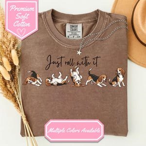 May include: A brown t-shirt featuring the text "Just roll with it" and a graphic of six beagles in different playful positions. The shirt has a crew neck and short sleeves. The text "Premium Soft Cotton" and "Multiple Colors Available" are also visible.