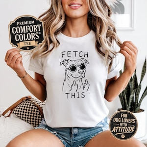 Peut inclure: T-shirt blanc avec un dessin noir d'un chien portant des lunettes de soleil et le texte "FETCH THIS". Le t-shirt porte une étiquette "Premium Comfort Colors Tee".