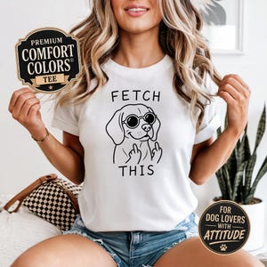 Peut inclure: T-shirt blanc avec un dessin noir d'un chien portant des lunettes de soleil et faisant un doigt d'honneur, avec le texte "FETCH THIS". Le t-shirt porte une étiquette "Premium Comfort Colors Tee".