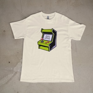 Puede incluir: Camiseta de color crema con un gráfico de juego de arcade retro. La máquina de arcade es verde y negra con un joystick y botones rojos. La pantalla muestra el texto "INSERT COIN TO CONTINUE".