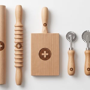 Può includere: Un set di utensili da cucina composto da un sottile bastoncino in legno, un mattarello liscio, un mattarello scanalato, una tavola per pasta rettangolare in legno e tre tagliapasta in metallo con manici in legno, ciascuno con il logo della croce svizzera.