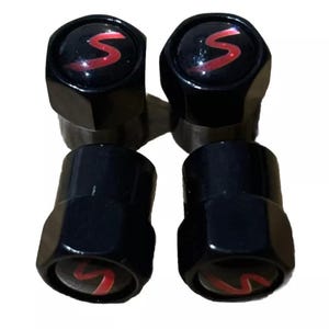 Puede incluir: Cuatro tapones negros para válvulas de neumáticos. Cada tapón tiene una base negra y una parte superior brillante y redondeada con un logotipo 'S' rojo. Los tapones están dispuestos en forma cuadrada, mostrando su diseño y acabado.
