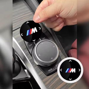 May include: Black and silver car interior control knob with a BMW logo. The logo is a circular design with the letters "M" and the BMW colors of blue, white, and red. The knob is being placed on a car's control panel.