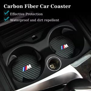 May include: Carbon fibre car coasters with the text "Carbon Fiber Car Coaster". The coasters are black with a carbon fibre pattern and the letters "IM" in white, blue, and red. The coasters are in a car's cup holders.