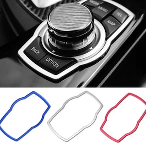 Könnte beinhalten: Nahaufnahme eines Infotainment-Bedienfelds eines Autos mit einem zentralen Drehregler und Tasten mit der Aufschrift "BACK" und "OPTION". Darunter drei farbige Zierteile: blau, silber und rot, zur Umrahmung des Bedienfelds.