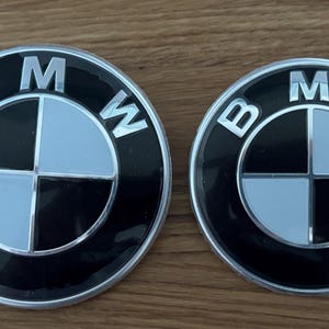 May include: Two circular BMW emblems with a black background and a silver border. The BMW logo is in the center, with a silver and black quadrant design and the letters "BMW" in silver.
