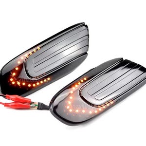 May include: Two black, oblong automotive side mirror turn signal lights with amber LED lights. Each light has a glossy black finish with a series of parallel lines on the top. Red and black wires with crocodile clips are attached.