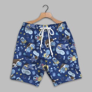 K&ouml;nnte beinhalten: Blaue Shorts mit einem durchgehenden Muster aus hellblauen, schildkr&ouml;tenartigen Wesen, gelben Seesternen, Ankern, Steuerr&auml;dern und kleinen wei&szlig;en Blasen. Die Shorts haben einen elastischen Bund mit wei&szlig;en Kordelz&uuml;gen und h&auml;ngen an einem Holzb&uuml;gel.