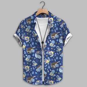 Puede incluir: Camisa azul abotonada con camiseta blanca y collar negro. La camisa presenta un patrón repetido de tortugas de dibujos animados, estrellas y elementos náuticos en tonos azules, marrones y amarillos. Las mangas están dobladas con tela blanca.