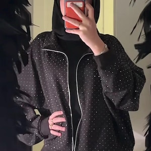 May include: Black zip-up jacket adorned with small, sparkling embellishments. The jacket features a white zip and is worn with a black turtleneck and trousers. The person in the image is wearing a black head covering.