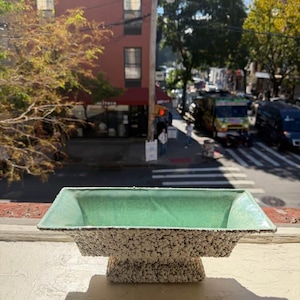 May include: A rectangular ceramic bowl with a smooth, light mint green interior and a textured, speckled black and white exterior. The bowl features a flared rim and sits on a sturdy, matching rectangular pedestal base, creating a footed design.